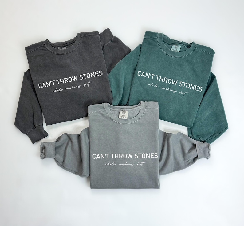 K&ouml;nnte beinhalten: Drei Sweatshirts in verschiedenen Farben, Grau, Schwarz und T&uuml;rkis, mit dem gleichen Text auf der Vorderseite: "CAN'T THROW STONES while washing feet".