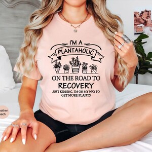 May include: A heather peach t-shirt featuring black text that reads I'M A PLANTAHOLIC ON THE ROAD TO RECOVERY JUST KIDDING, I'M ON MY WAY TO GET MORE PLANTS, accompanied by a graphic illustration of five potted plants in a row.