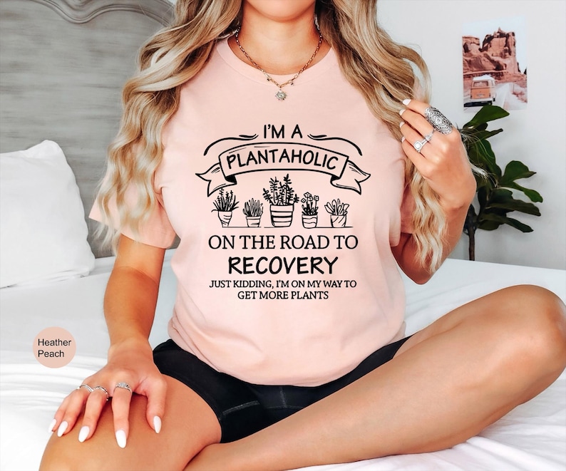 May include: A heather peach t-shirt featuring black text that reads I'M A PLANTAHOLIC ON THE ROAD TO RECOVERY JUST KIDDING, I'M ON MY WAY TO GET MORE PLANTS, accompanied by a graphic illustration of five potted plants in a row.