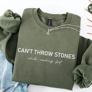 May include: A military green crewneck sweatshirt featuring the white text CAN'T THROW STONES while washing feet printed on the front. The garment has long sleeves with ribbed cuffs and a round neckline, displayed with a silver chain necklace.