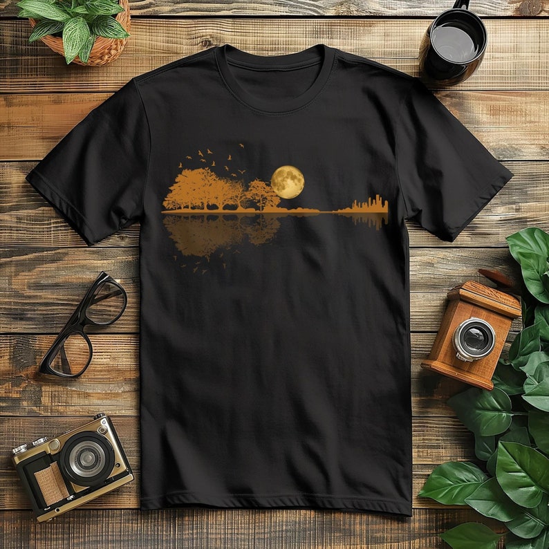 Pode incluir: Camiseta preta com um design em forma de guitarra que apresenta uma lua cheia, &aacute;rvores e p&aacute;ssaros em tons de ouro e castanho. O design evoca uma cena natural. A camiseta &eacute; exibida em uma superf&iacute;cie de madeira.