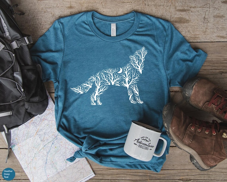 May include: Teal t-shirt featuring a white wolf design made of tree branches and a crescent moon. A white mug with the text "And so the adventure begins" sits next to the shirt. Hiking boots and a backpack are also in the image.