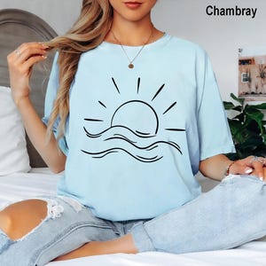 Pu&ograve; includere: Una t-shirt a maniche corte color azzurro con una grafica a linee nere raffigurante un sole che sorge sopra le onde dell'oceano sul petto. Il capo &egrave; etichettato come Chambray ed &egrave; abbinato a jeans azzurri con ampi strappi sulle ginocchia.