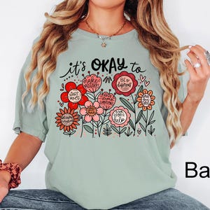 May include: A sage green short-sleeve t-shirt featuring a graphic of colorful flowers with text that reads it's OKAY to make mistakes, be different, start over, have strong emotions, have bad days, ask for help, and not know everything.