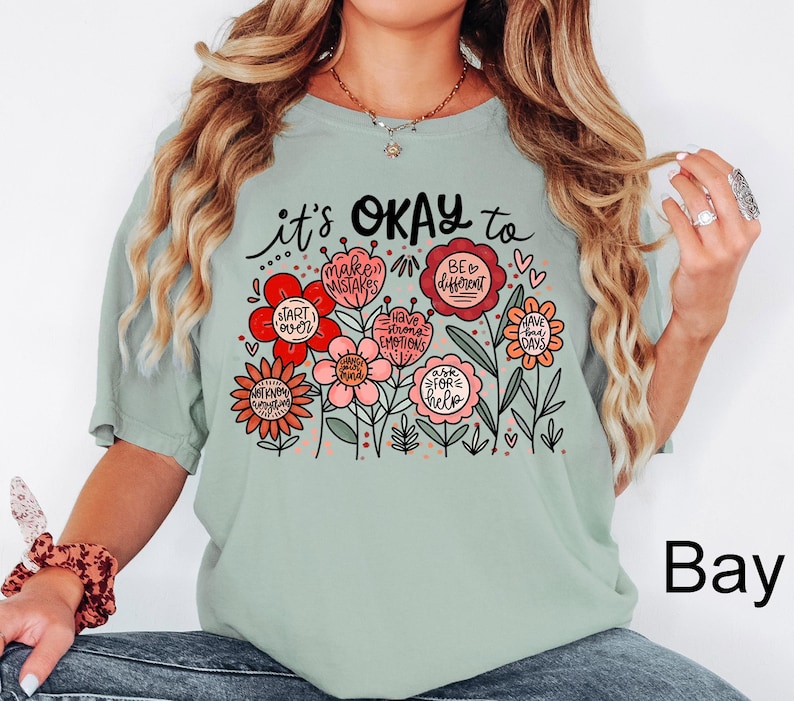 May include: A sage green short-sleeve t-shirt featuring a graphic of colorful flowers with text that reads it's OKAY to make mistakes, be different, start over, have strong emotions, have bad days, ask for help, and not know everything.