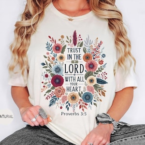 May include: Cream-colored t-shirt featuring a floral design with the text "TRUST IN THE LORD WITH ALL YOUR HEART" and "Proverbs 3:5". The design includes colorful flowers and foliage.