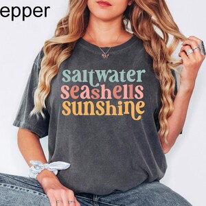 K&ouml;nnte beinhalten: Dunkelgraues T-Shirt mit den Worten "saltwater seashells sunshine" in T&uuml;rkis, Koralle und Gelb. Das Shirt hat eine lockere Passform und kurze &Auml;rmel. Das Wort "Pepper" befindet sich oben links.