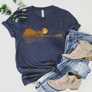 Pode incluir: Uma camiseta azul marinho com um gr&aacute;fico de uma silhueta de guitarra refletindo uma paisagem com uma lua cheia. A camisa &eacute; combinada com jeans azuis e botinhas bege. A camisa &eacute; feita de um material macio.