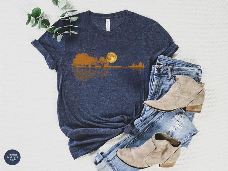 Pode incluir: Uma camiseta azul marinho com um gr&aacute;fico de uma silhueta de guitarra refletindo uma paisagem com uma lua cheia. A camisa &eacute; combinada com jeans azuis e botinhas bege. A camisa &eacute; feita de um material macio.