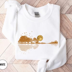 Pode incluir: Camisola branca com um design de paisagem. Apresenta uma lua dourada, &aacute;rvores e um horizonte da cidade refletido na &aacute;gua, com p&aacute;ssaros voando por cima. A palavra "WHITE" est&aacute; num c&iacute;rculo branco na parte inferior.