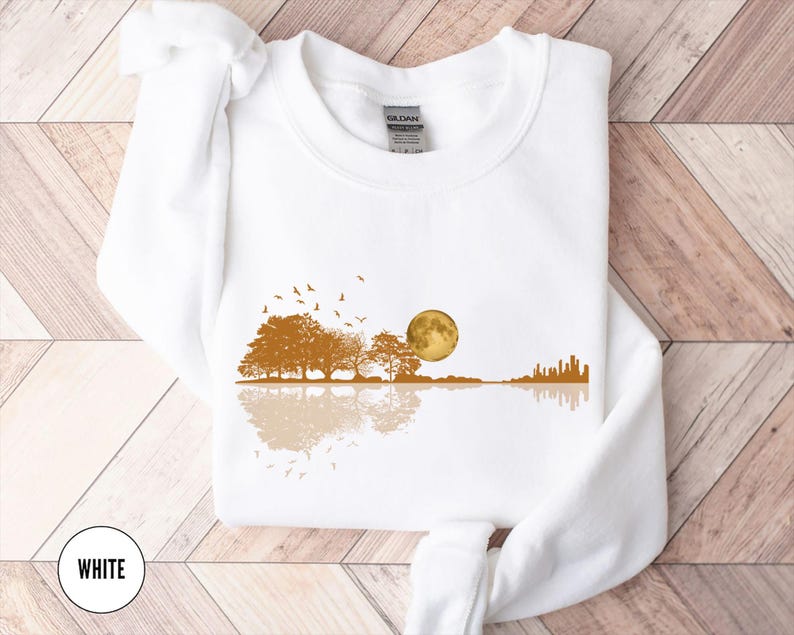 Pode incluir: Camisola branca com um design de paisagem. Apresenta uma lua dourada, &aacute;rvores e um horizonte da cidade refletido na &aacute;gua, com p&aacute;ssaros voando por cima. A palavra "WHITE" est&aacute; num c&iacute;rculo branco na parte inferior.