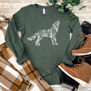 May include: A dark green long-sleeve shirt with a white wolf design made of tree branches and a crescent moon. The shirt is paired with a brown and white plaid shirt, black jeans, and brown boots.