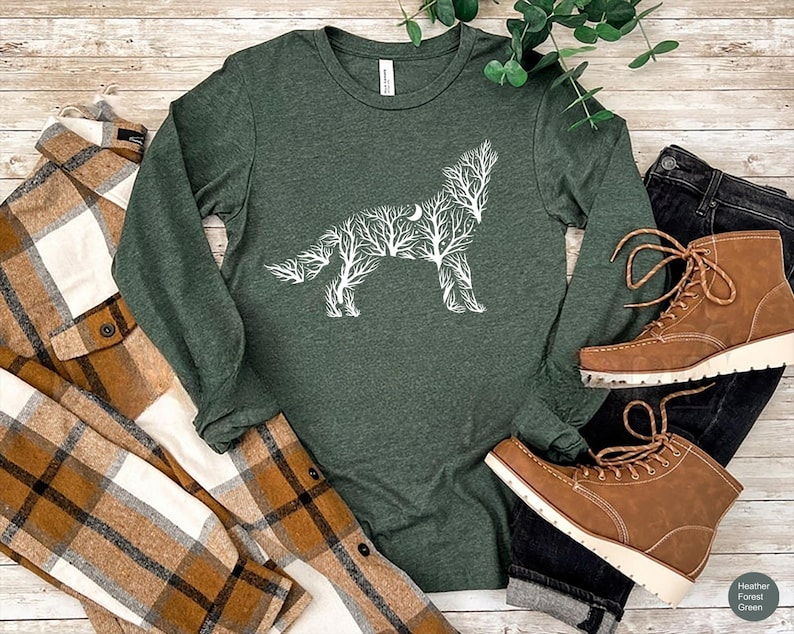 May include: A dark green long-sleeve shirt with a white wolf design made of tree branches and a crescent moon. The shirt is paired with a brown and white plaid shirt, black jeans, and brown boots.