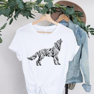 May include: White t-shirt featuring a black silhouette of a howling wolf, composed of tree branches and a crescent moon. The shirt is displayed on a wooden hanger with a denim jacket and a hat in the background. The design is a nature-inspired graphic.