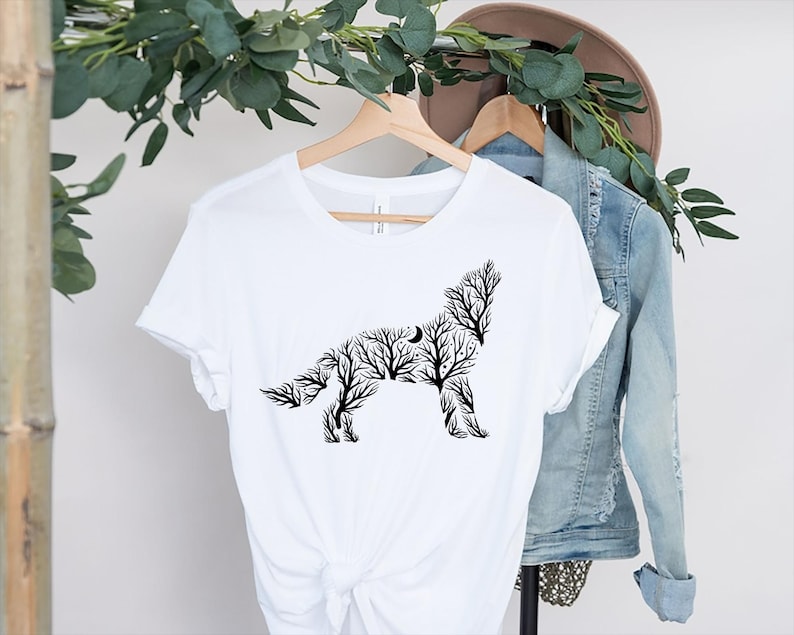 May include: White t-shirt featuring a black silhouette of a howling wolf, composed of tree branches and a crescent moon. The shirt is displayed on a wooden hanger with a denim jacket and a hat in the background. The design is a nature-inspired graphic.