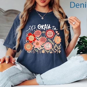 May include: A dark blue short-sleeve t-shirt featuring a graphic of colorful flowers with text that reads it's OKAY to, START OVER, MAKE MISTAKES, BE DIFFERENT, HAVE STRONG EMOTIONS, HAVE BAD DAYS, ASK FOR HELP, and NOT KNOW EVERYTHING.