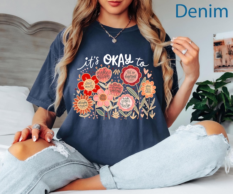 May include: A dark blue short-sleeve t-shirt featuring a graphic of colorful flowers with text that reads it's OKAY to, START OVER, MAKE MISTAKES, BE DIFFERENT, HAVE STRONG EMOTIONS, HAVE BAD DAYS, ASK FOR HELP, and NOT KNOW EVERYTHING.