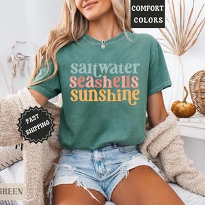 K&ouml;nnte beinhalten: Ein hellgr&uuml;nes T-Shirt mit den Worten "saltwater seashells sunshine" in einer bunten Retro-Schrift. Das Shirt wird mit Jeansshorts und einer cremefarbenen Strickjacke kombiniert. Das Bild hat eine sommerliche, strandige Atmosph&auml;re.