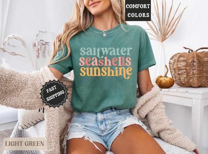 K&ouml;nnte beinhalten: Ein hellgr&uuml;nes T-Shirt mit den Worten "saltwater seashells sunshine" in einer bunten Retro-Schrift. Das Shirt wird mit Jeansshorts und einer cremefarbenen Strickjacke kombiniert. Das Bild hat eine sommerliche, strandige Atmosph&auml;re.