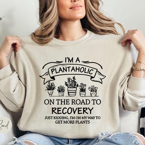 May include: A sand-colored crewneck sweatshirt featuring black text that reads I'M A PLANTAHOLIC ON THE ROAD TO RECOVERY JUST KIDDING, I'M ON MY WAY TO GET MORE PLANTS, accompanied by a graphic illustration of five potted plants in a row.
