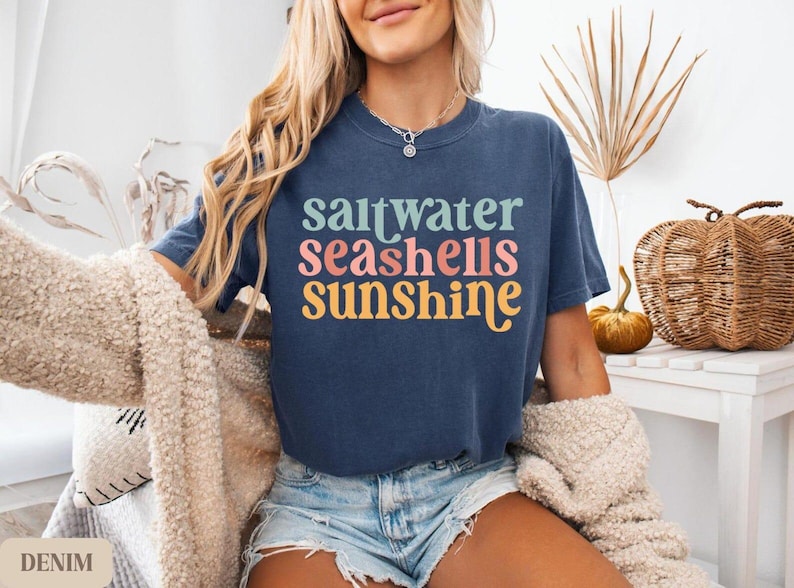 K&ouml;nnte beinhalten: Ein marineblaues T-Shirt mit den Worten "saltwater seashells sunshine" in Pastellfarben. Das Model tr&auml;gt Jeansshorts und eine cremefarbene Strickjacke. Ein dekorativer K&uuml;rbis und ein Weidenkorb befinden sich im Hintergrund.