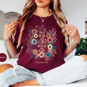May include: Maroon t-shirt featuring a floral design with the text "Trust in the Lord with all your heart" and "Proverbs 3:5". The design includes colorful flowers and leaves. The shirt is a crew neck.