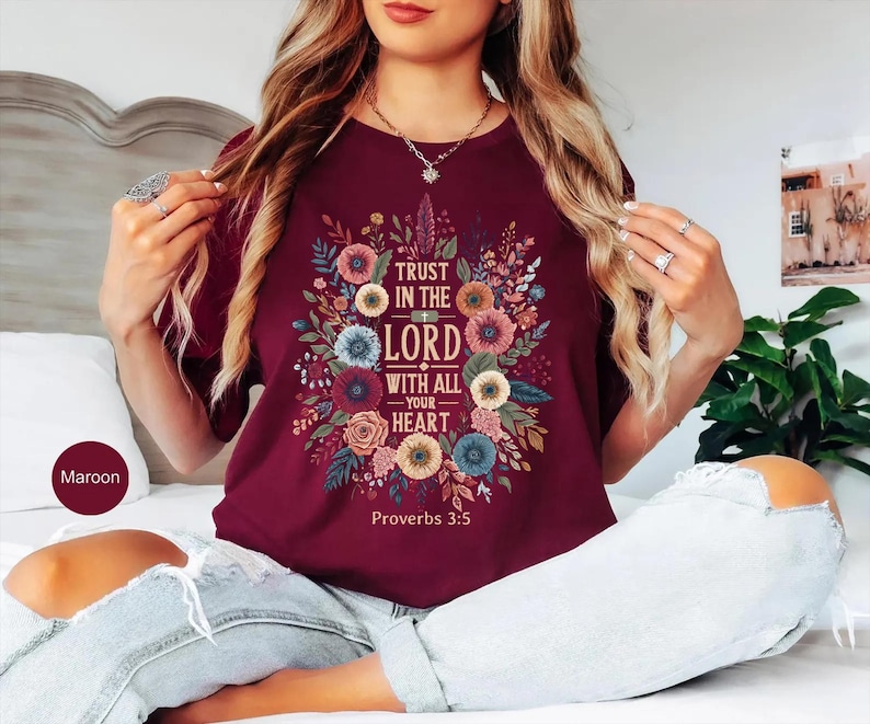 May include: Maroon t-shirt featuring a floral design with the text "Trust in the Lord with all your heart" and "Proverbs 3:5". The design includes colorful flowers and leaves. The shirt is a crew neck.