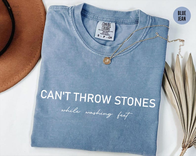 K&ouml;nnte beinhalten: Ein T-Shirt aus Jeansblau mit dem Aufdruck "CAN'T THROW STONES while washing feet" in Wei&szlig; auf der Vorderseite.