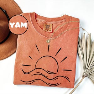 Pu&ograve; includere: Una t-shirt a maniche corte color terracotta con una grafica a linee nere che raffigura un sole che sorge sopra le onde dell'oceano. La maglia presenta un girocollo e un'etichetta Comfort Colors. Un inserto circolare mostra il testo YAM.