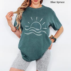 Pu&ograve; includere: Una t-shirt a maniche corte color abete blu con una grafica a linee bianche che raffigura un sole che sorge sopra le onde dell'oceano. Il capo presenta un girocollo e una vestibilit&agrave; rilassata. Il testo Blue Spruce &egrave; visibile nell'immagine.