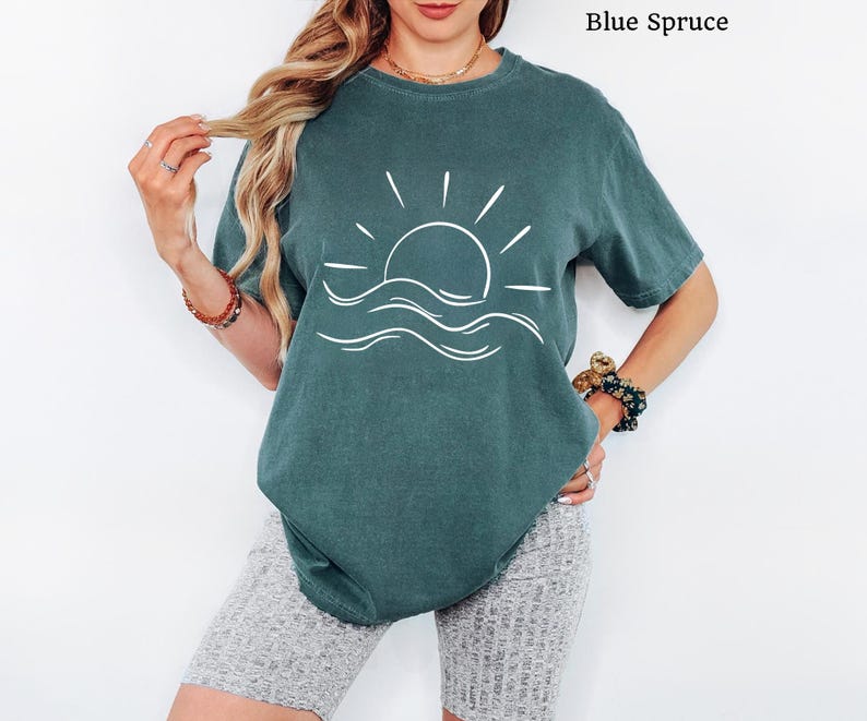 Pu&ograve; includere: Una t-shirt a maniche corte color abete blu con una grafica a linee bianche che raffigura un sole che sorge sopra le onde dell'oceano. Il capo presenta un girocollo e una vestibilit&agrave; rilassata. Il testo Blue Spruce &egrave; visibile nell'immagine.