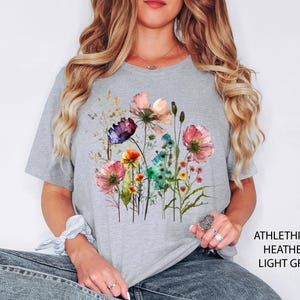 May include: Light grey t-shirt featuring a watercolor floral design with pink, purple, orange, and blue flowers. The shirt has the text "ATHLETHICH HEATHER LIGHT GREY" printed on the side.