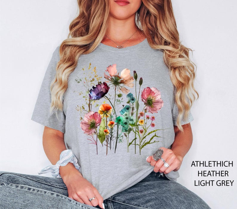 May include: Light grey t-shirt featuring a watercolor floral design with pink, purple, orange, and blue flowers. The shirt has the text "ATHLETHICH HEATHER LIGHT GREY" printed on the side.