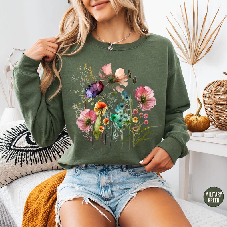 May include: Olive green sweatshirt featuring a watercolor floral design with pink, purple, blue, and yellow flowers. The sweatshirt has long sleeves and a crew neck. The words "MILITARY GREEN" are in a small circle on the bottom right.