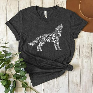 May include: Dark heather gray t-shirt featuring a white wolf design made of tree branches and a crescent moon. The t-shirt is knotted at the bottom and has a crew neck. The shirt is displayed on a light wood surface.