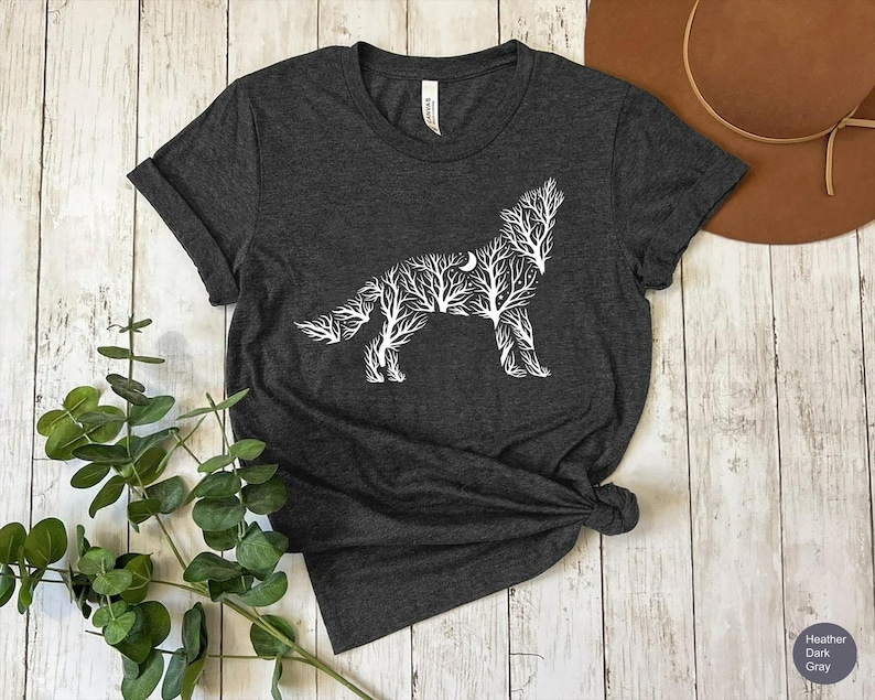 May include: Dark heather gray t-shirt featuring a white wolf design made of tree branches and a crescent moon. The t-shirt is knotted at the bottom and has a crew neck. The shirt is displayed on a light wood surface.