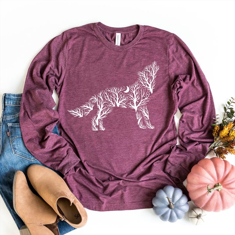 May include: Burgundy long-sleeve shirt featuring a white wolf design made of tree branches and a crescent moon. The shirt is displayed with blue jeans, brown shoes, and decorative pumpkins, suggesting a fall theme.