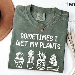 May include: Hemp-colored t-shirt with the phrase "SOMETIMES I WET MY PLANTS" in white. Below the text are four line-drawn potted plants. A gold necklace with a pendant rests on the shirt. A brown hat is partially visible.