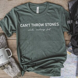 May include: A heather forest green short-sleeve t-shirt featuring the white text CAN'T THROW STONES while washing feet printed on the front. The shirt has a crew neckline and a standard fit, showcasing a soft, textured fabric appearance.