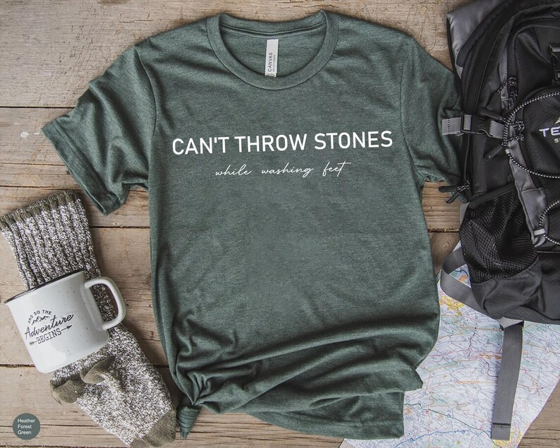 May include: A heather forest green short-sleeve t-shirt featuring the white text CAN'T THROW STONES while washing feet printed on the front. The shirt has a crew neckline and a standard fit, showcasing a soft, textured fabric appearance.
