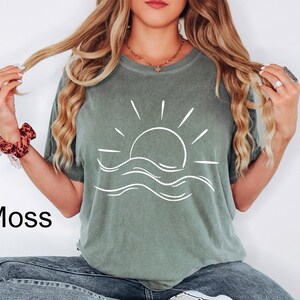 Pu&ograve; includere: Una t-shirt a maniche corte color verde muschio con una grafica a linee bianche raffigurante un sole che sorge sopra le onde dell'oceano al centro. La maglia ha un girocollo e una vestibilit&agrave; ampia, con la parola Moss stampata in nero in basso.