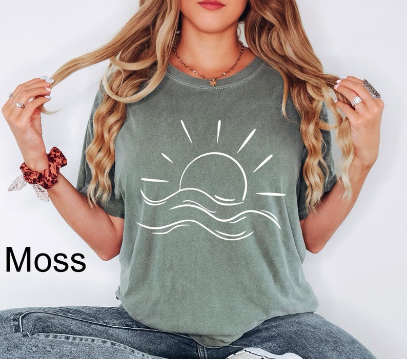 Pu&ograve; includere: Una t-shirt a maniche corte color verde muschio con una grafica a linee bianche raffigurante un sole che sorge sopra le onde dell'oceano al centro. La maglia ha un girocollo e una vestibilit&agrave; ampia, con la parola Moss stampata in nero in basso.