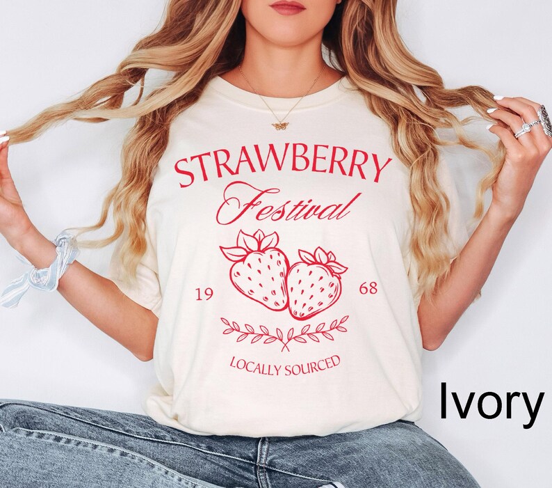 Op de afbeelding: Een ivoorkleurig t-shirt met rode grafische tekst die STRAWBERRY Festival, de cijfers 19 en 68, en de woorden LOCALLY SOURCED weergeeft. Het ontwerp bevat een centrale illustratie van twee aardbeien met bladeren en een decoratief rankelement.