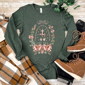 May include: A dark green long-sleeve shirt with a floral and cross design. The design includes the text "Pray for us who have recourse to thee". Brown boots and a plaid shirt are also in the image.