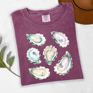 May include: A maroon Comfort Colors t-shirt featuring a watercolor design of six oyster shells. The shells are in shades of white, blue, and pink. The shirt is folded and has a tag in the upper right corner.