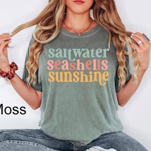 K&ouml;nnte beinhalten: Moosgr&uuml;nes T-Shirt mit dem Schriftzug "saltwater seashells sunshine" in bunten Farben. Das Shirt hat eine lockere Passform und kurze &Auml;rmel. Das Wort "Moss" ist unten links in Schwarz aufgedruckt.