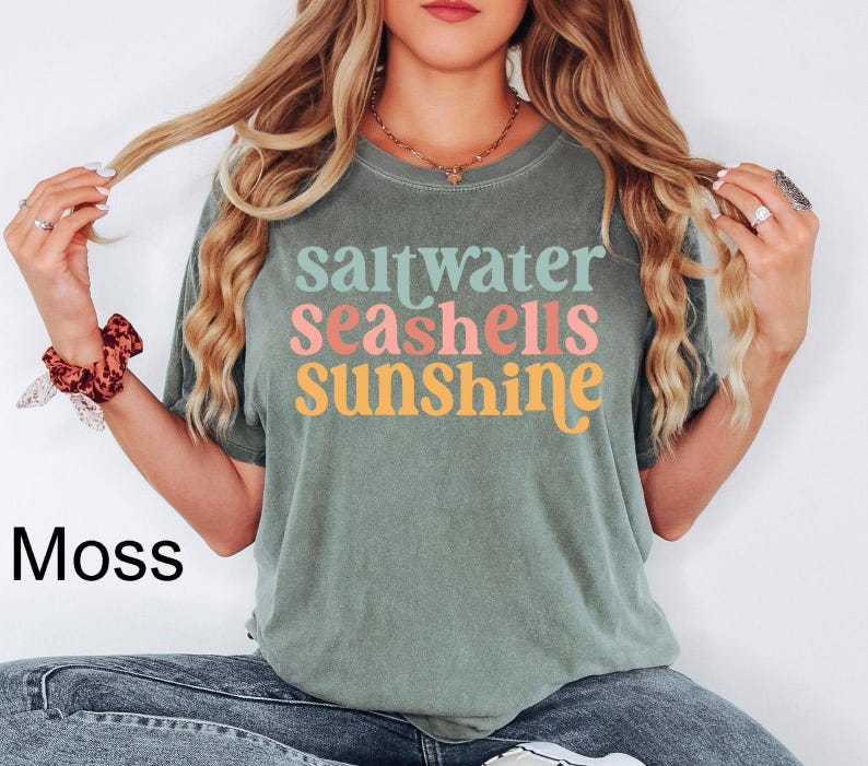 K&ouml;nnte beinhalten: Moosgr&uuml;nes T-Shirt mit dem Schriftzug "saltwater seashells sunshine" in bunten Farben. Das Shirt hat eine lockere Passform und kurze &Auml;rmel. Das Wort "Moss" ist unten links in Schwarz aufgedruckt.