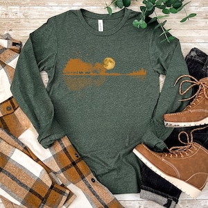 Pode incluir: Uma camisa de manga comprida verde escuro com um design de paisagem com lua cheia, &aacute;rvores e uma silhueta de guitarra. A camisa &eacute; combinada com botas castanhas, uma camisa xadrez e jeans escuros. A camisa tem a inscri&ccedil;&atilde;o "Heather Forest Green".