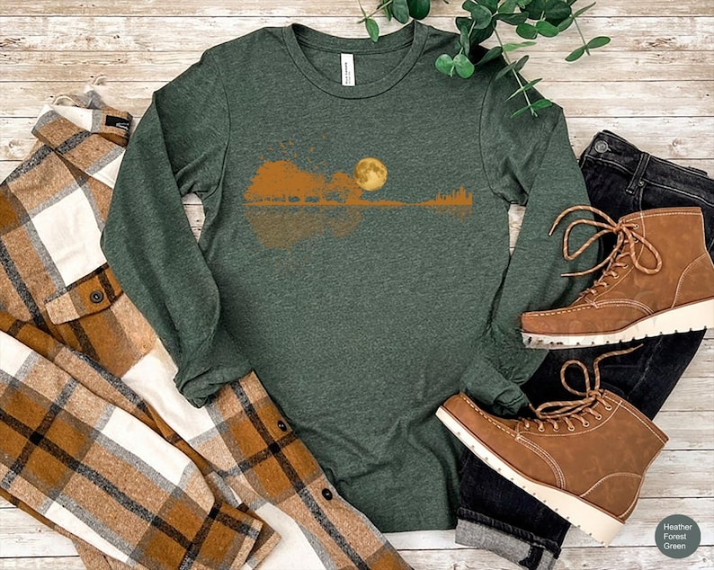 Pode incluir: Uma camisa de manga comprida verde escuro com um design de paisagem com lua cheia, &aacute;rvores e uma silhueta de guitarra. A camisa &eacute; combinada com botas castanhas, uma camisa xadrez e jeans escuros. A camisa tem a inscri&ccedil;&atilde;o "Heather Forest Green".