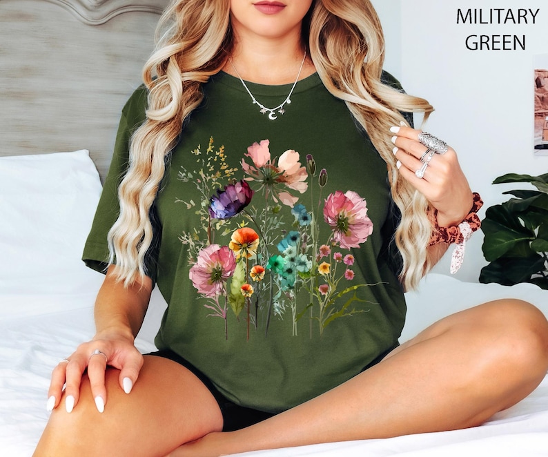 May include: Olive green t-shirt featuring a watercolor floral design with pink, purple, orange, and blue flowers. The text "MILITARY GREEN" is printed in the upper right corner. The shirt is a crew neck.
